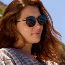 on model - female at the beach wearing dime optics featuring the silverlake round sunglasses with a black frame and grey lenses angled view